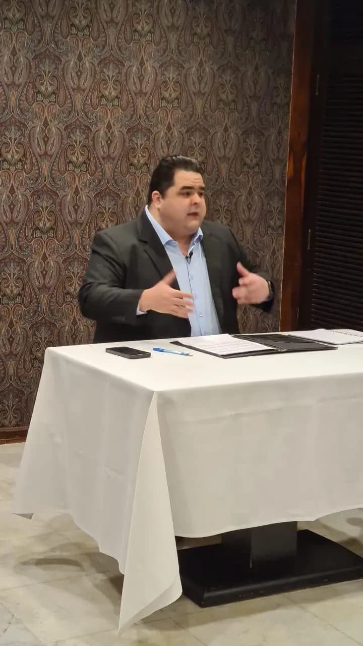 Mateus Gomes falando sentado a uma mesa durante uma apresentação
